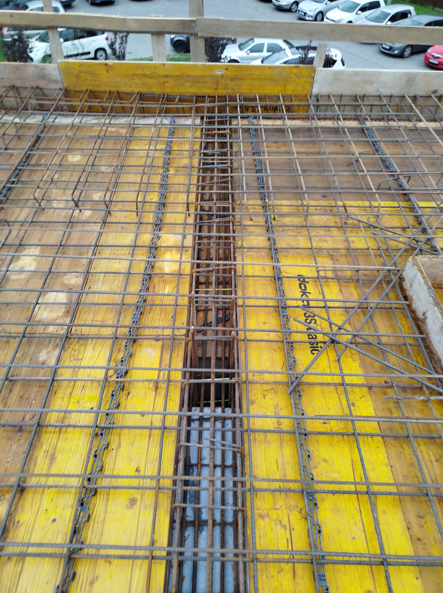 Zbrojenie stropu na żółtym szalunku z widocznymi rurami instalacyjnymi w kanale. Siatka z prętów zbrojeniowych ułożona regularnie. Widok z góry na plac budowy.