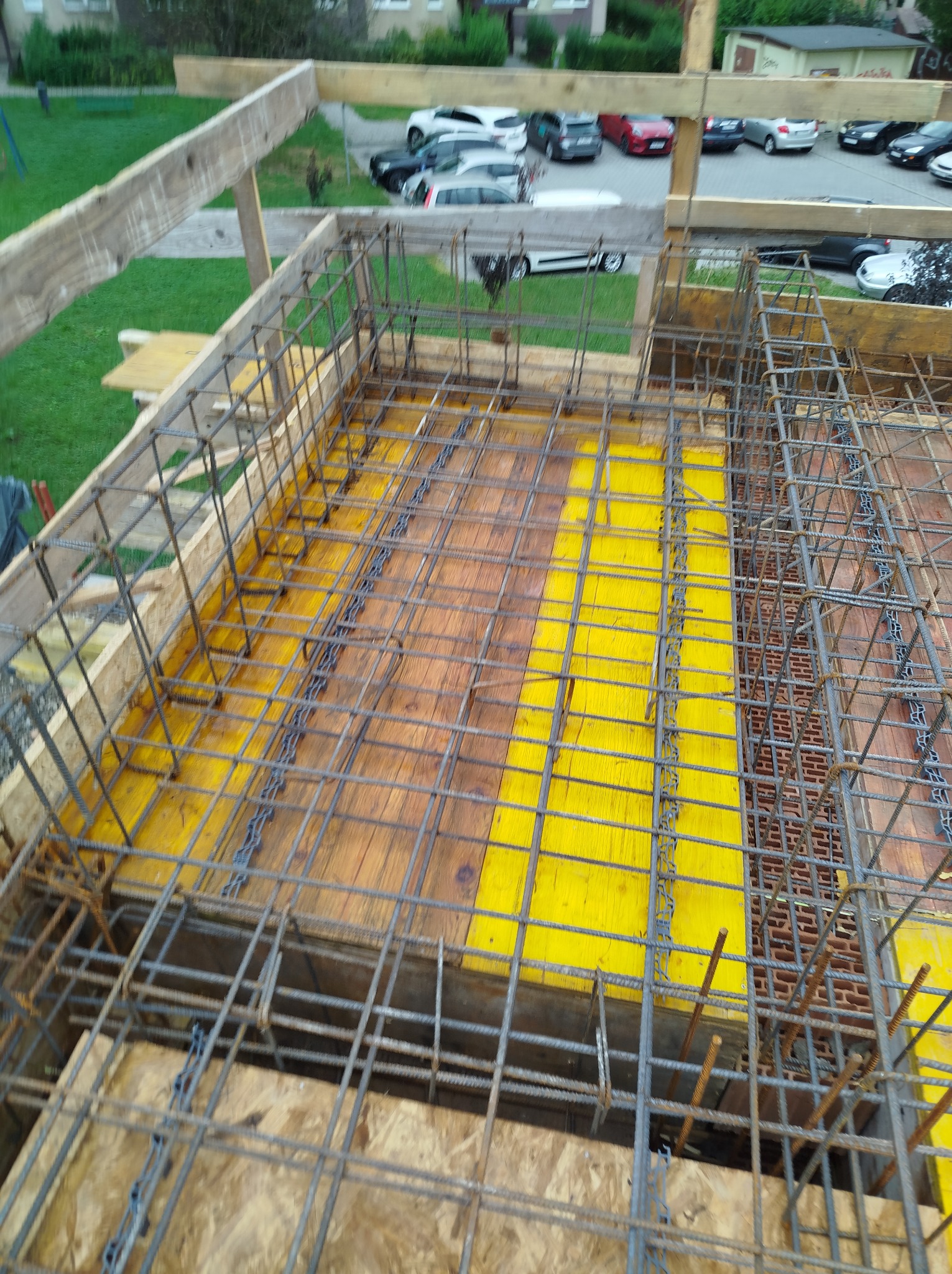 Zbrojenie fundamentu z żółtymi szalunkami i widoczną konstrukcją drewnianą. W tle zieleń i zaparkowane samochody. Kąt ujęcia z góry.