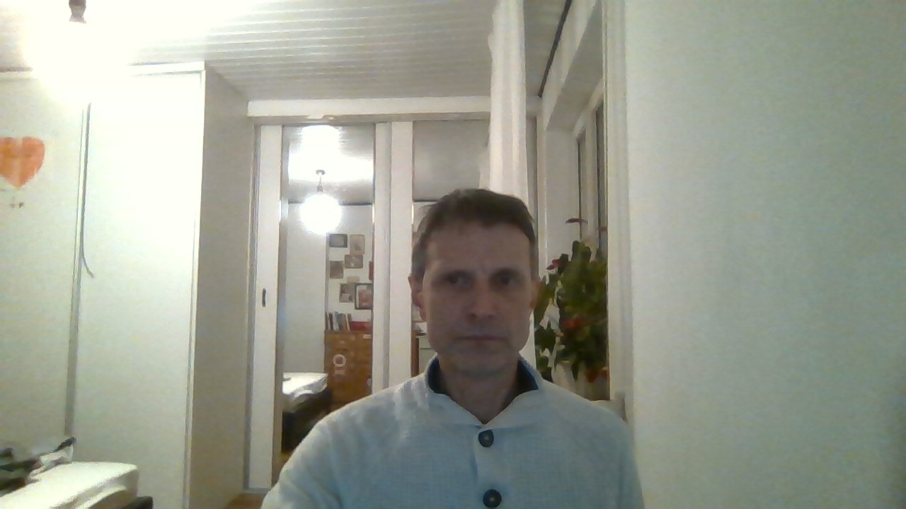 Mężczyzna w jasnym swetrze na tle wnętrza z szafą, oknem i dekoracjami. Ujęcie z bliska, oświetlenie mieszane, widoczne elementy wyposażenia.