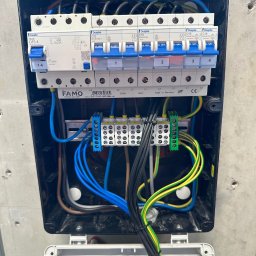 MaxMed Elektro - Rozdzielnica elektryczna w czarnej obudowie z widocznymi bezpiecznikami, przewodami w kolorach niebieskim, żółto-zielonym i czarnym. Widoczne oznaczenia producenta Famo i Doepke.