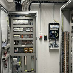 MaxMed Elektro - Rozdzielnia elektryczna z otwartymi drzwiami, widoczne liczne kable, bezpieczniki i elementy sterujące. Na ścianie dodatkowa skrzynka z instalacją.