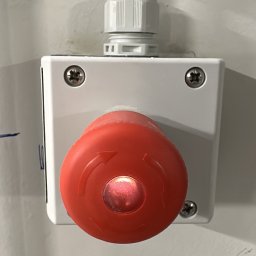 MaxMed Elektro - Czerwony przycisk bezpieczeństwa z białym korpusem, zamontowany na ścianie z widocznym przewodem. Przycisk oznaczony strzałkami. Widoczne śruby mocujące.