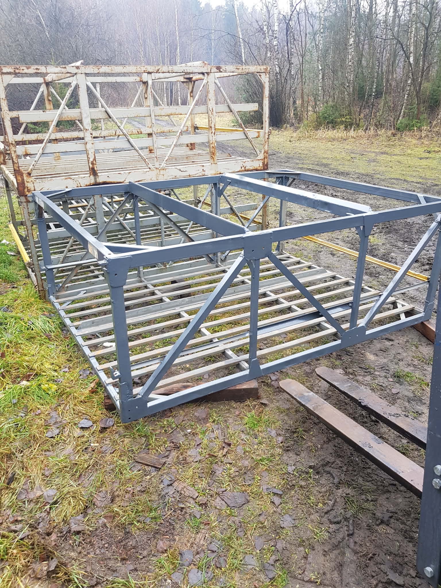 Konstrukcja metalowa z kratownicą, pomalowana na szaro, leżąca na trawie. Widoczne elementy wózka widłowego. Tło stanowią drzewa bez liści.