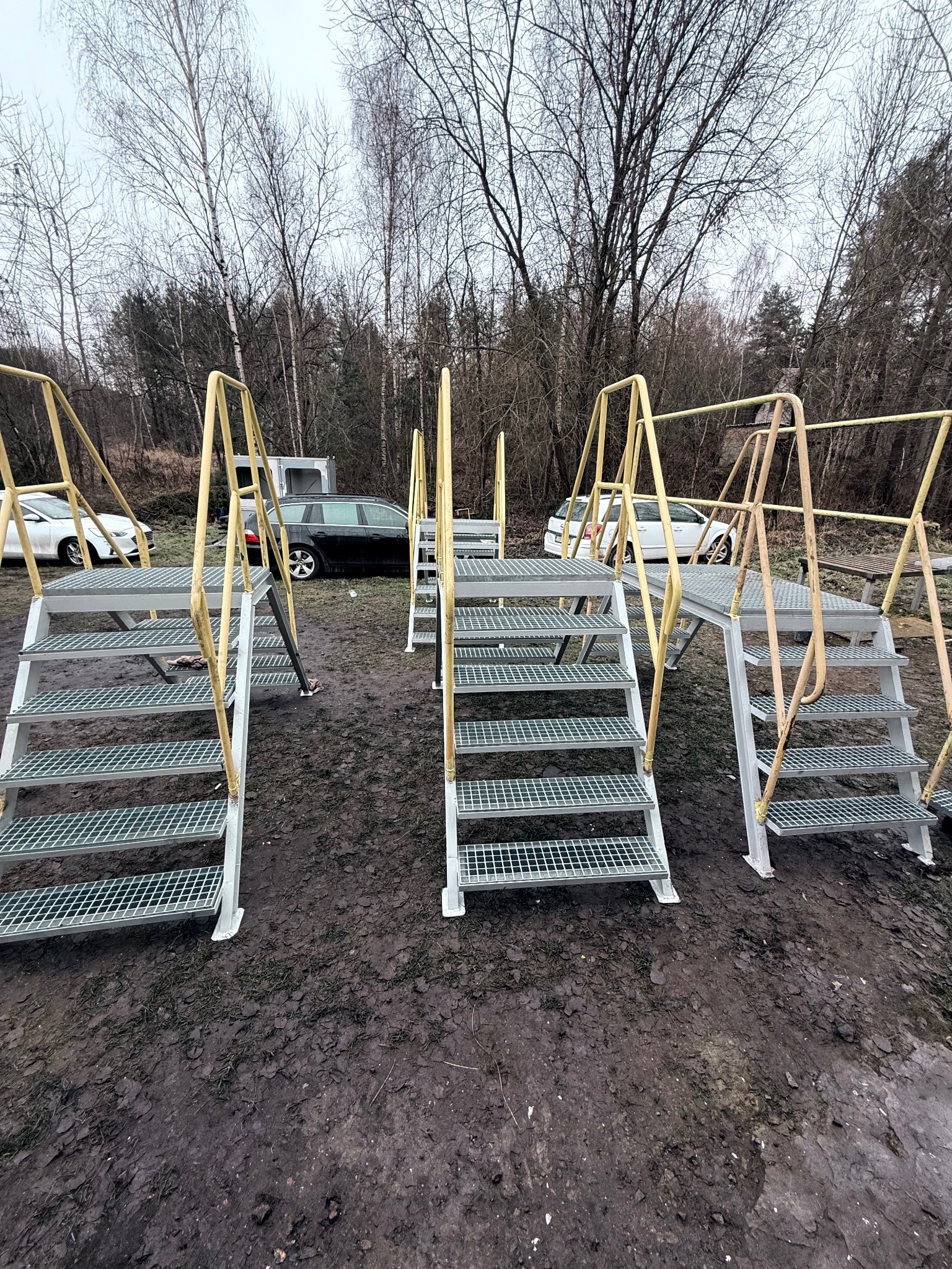 Metalowe schody z żółtymi poręczami na zewnątrz, z widocznymi samochodami w tle, prezentujące konstrukcje metalowe.