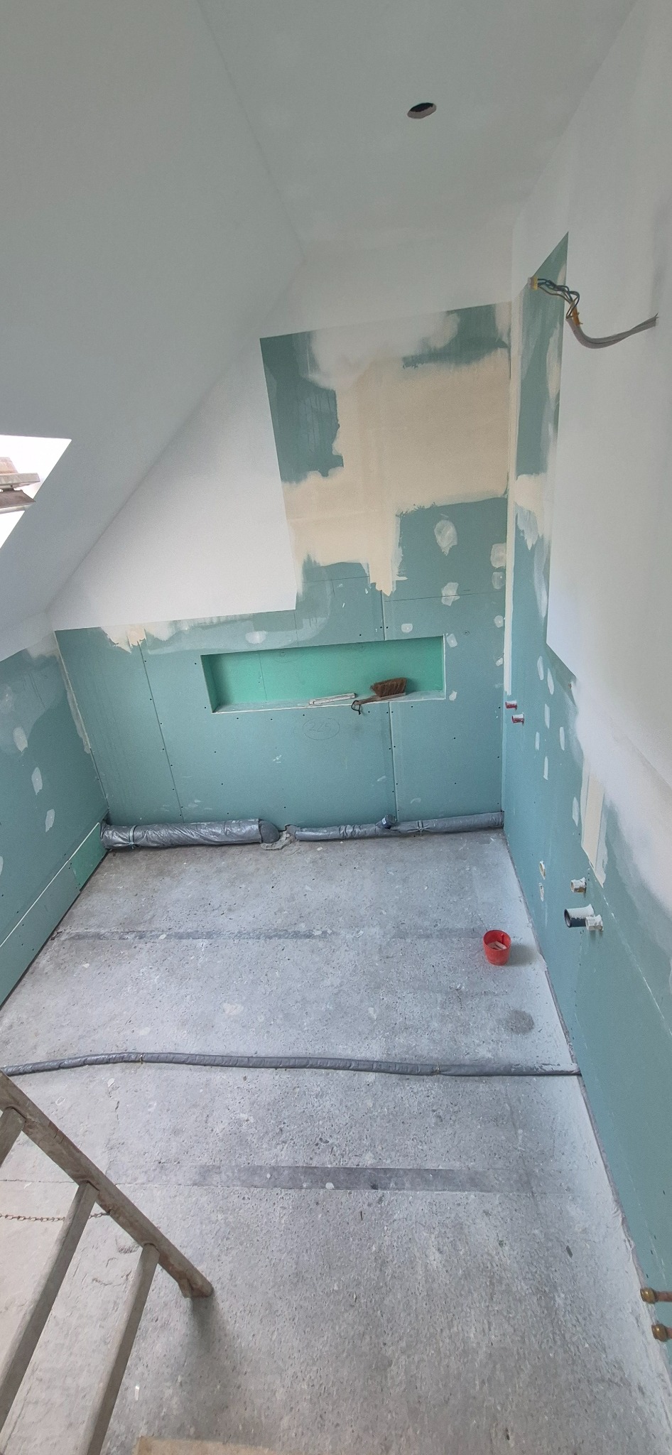 Pokój na poddaszu w trakcie prac: ściany z płyt gipsowo-kartonowych, częściowo zagruntowane, widoczne instalacje. Wnęka w ścianie, drabina w rogu.