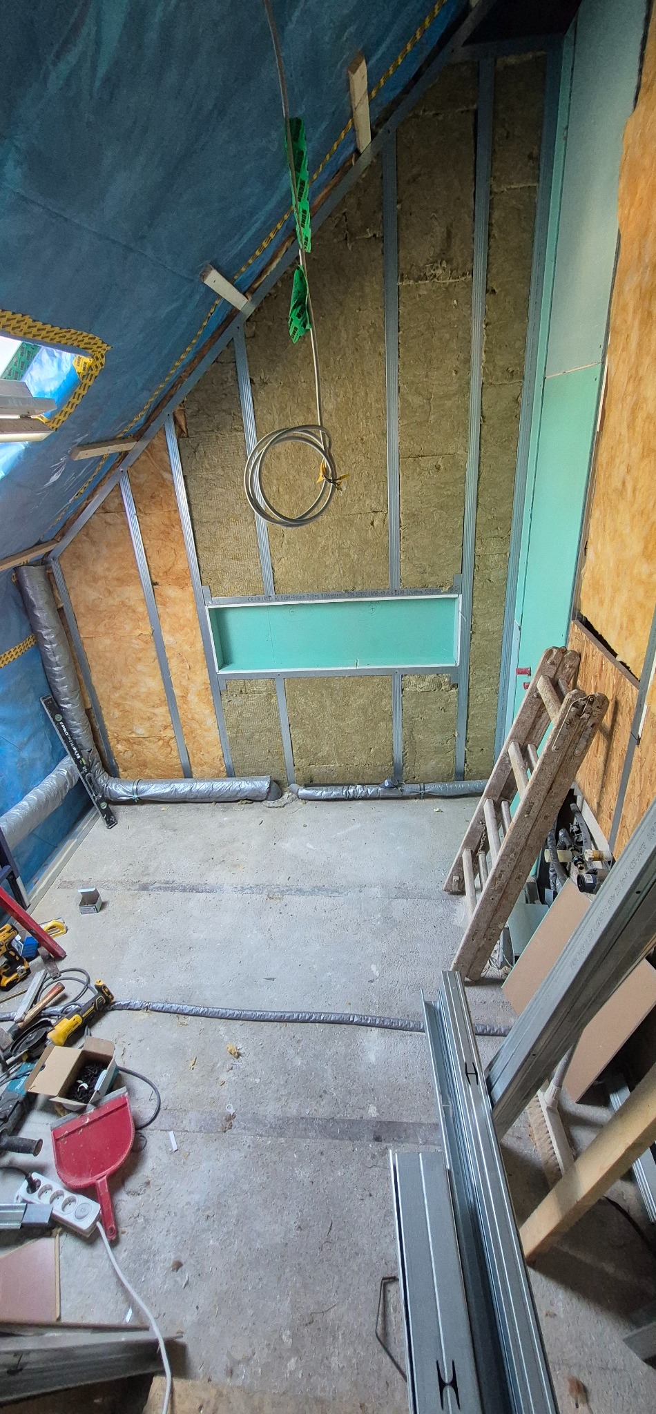 Montaż izolacji w poddaszu: wełna mineralna między metalowymi profilami, widoczne elementy konstrukcji, narzędzia na podłodze, fragment okna dachowego.