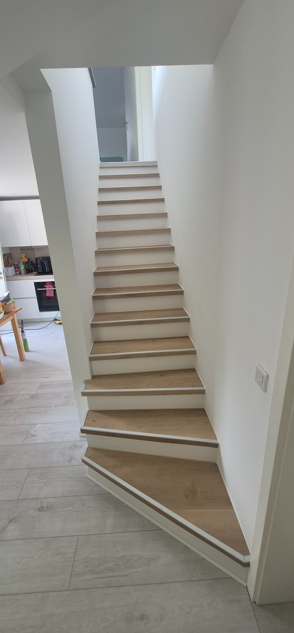 Nowoczesne schody z drewnopodobnymi panelami i białymi podstopnicami, prowadzące na górę. Widoczne wykończenie podłogi w podobnym stylu.