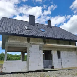 Surowy dom z białych bloczków, czarny dach z oknami dachowymi i kominami, widok z dołu na tle błękitnego nieba z chmurami. Stan surowy otwarty.