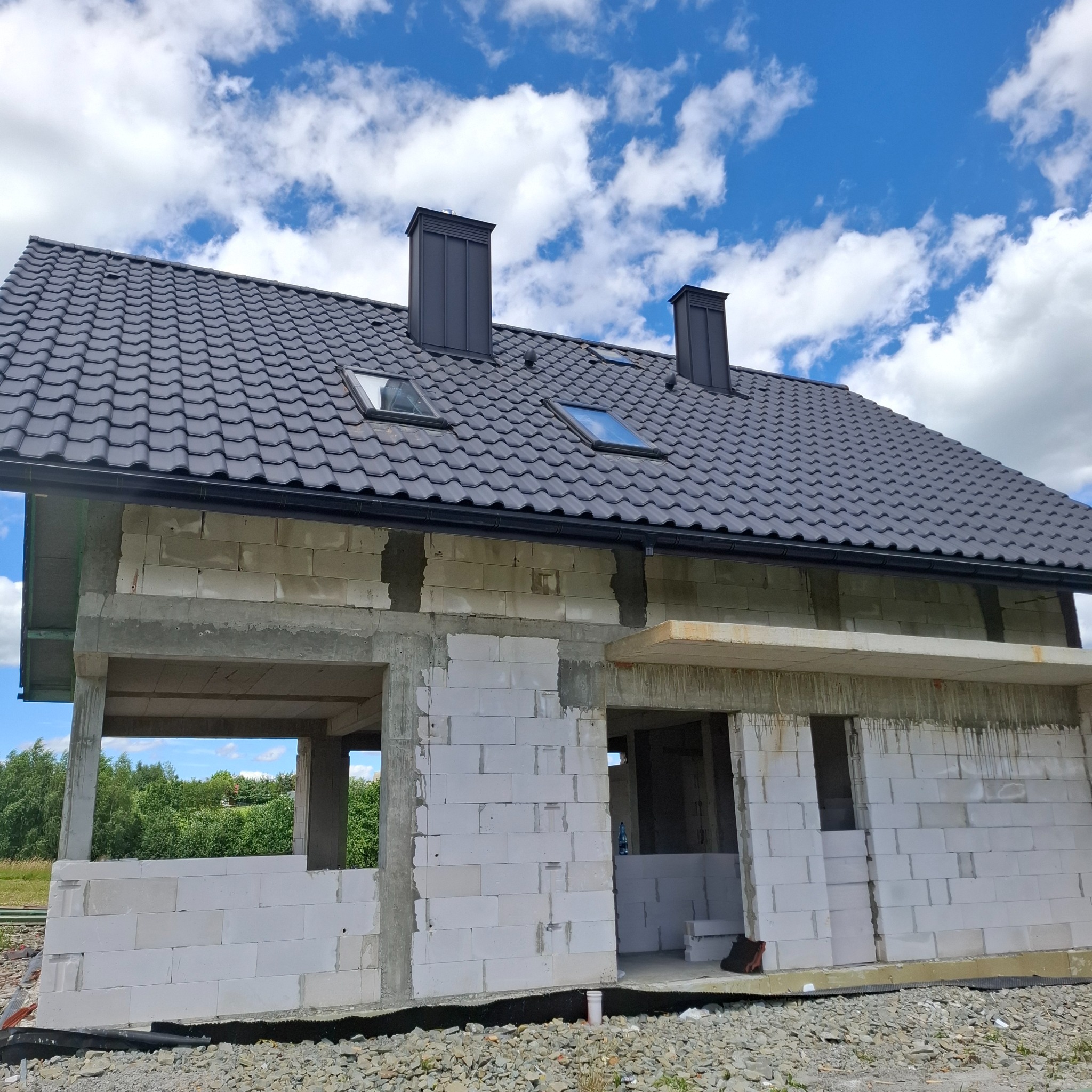 Surowy dom z białych bloczków, czarny dach z oknami dachowymi i kominami, widok z dołu na tle błękitnego nieba z chmurami. Stan surowy otwarty.