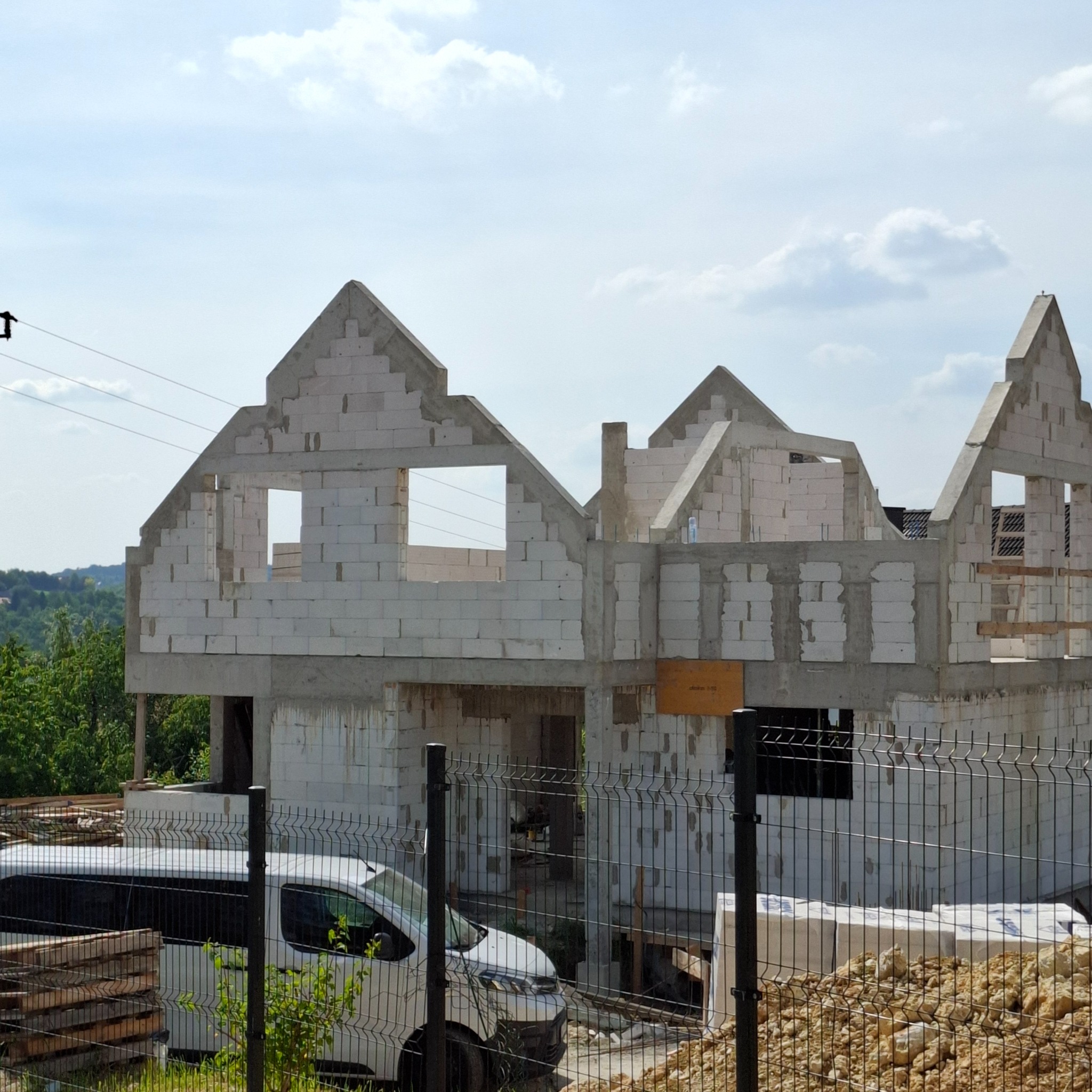 Wznoszenie domu z białych bloczków, widoczne otwory okienne i zarys dachu. Na pierwszym planie ogrodzenie i zaparkowany biały bus. Budowa domu w trakcie realizacji.