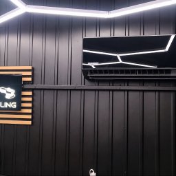 KMS-PRO ADAM DĄBROWSKI - Czarny klimatyzator Rotenso z białymi liniami LED na ścianie z blachy trapezowej w warsztacie autodetailingu, oświetlony lampą LED. Widoczny znak świetlny firmy.