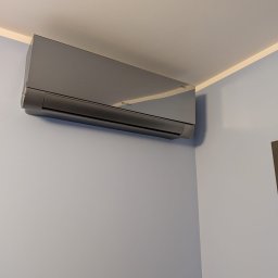 KMS-PRO ADAM DĄBROWSKI - Szary klimatyzator ROTENSO zamontowany na jasnoniebieskiej ścianie w rogu pokoju, widoczny fragment ramy drzwi i okna. Nowoczesny design, minimalistyczny styl.