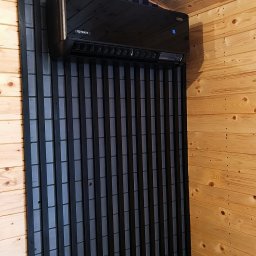 KMS-PRO ADAM DĄBROWSKI - Czarny klimatyzator Rotenso na tle dekoracyjnej, pionowej boazerii w pomieszczeniu wykończonym drewnem. Widoczny wyświetlacz temperatury.