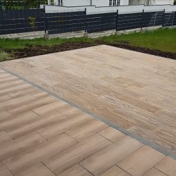 INT - Wykończony taras z kostki brukowej imitującej drewno, z szarą obwódką, na tle domu z ciemnym dachem i czarnym ogrodzeniem, w słoneczny dzień.