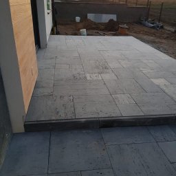 INT - Ułożona kostka granitowa w odcieniach szarości, tworząca taras przy budynku. W tle widoczne prace ziemne i ogrodzenie.