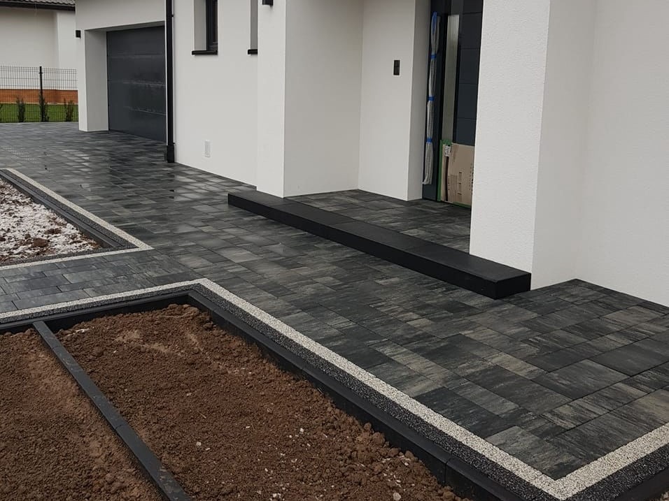 Nowoczesna, grafitowa kostka brukowa z obramowaniem z jasnego grysu, tworząca ścieżkę i rabaty przed domem z białą elewacją i czarną bramą garażową.