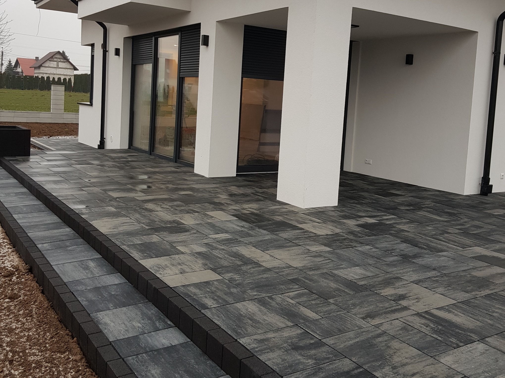 Nowoczesny taras z szarej kostki brukowej ułożonej we wzór jodełki, z czarnymi obrzeżami i schodami, przylegający do białego domu z dużymi oknami.