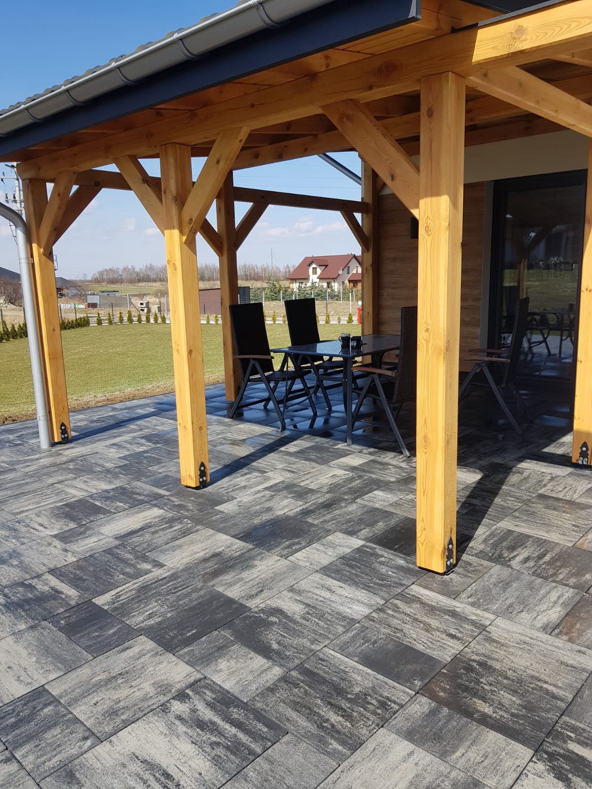 Zadaszony taras z kostki brukowej w odcieniach szarości i czerni. Widoczne drewniane słupy i meble ogrodowe. W tle dom i zielony trawnik.