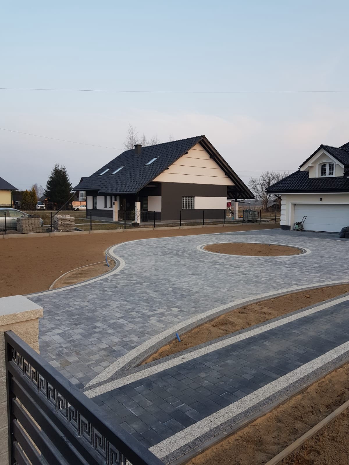 Nowoczesny podjazd z szarej kostki brukowej ułożonej we wzór, z obrzeżami z jasnego kamienia. W tle domy z ciemnymi dachami i czarnym ogrodzeniem.