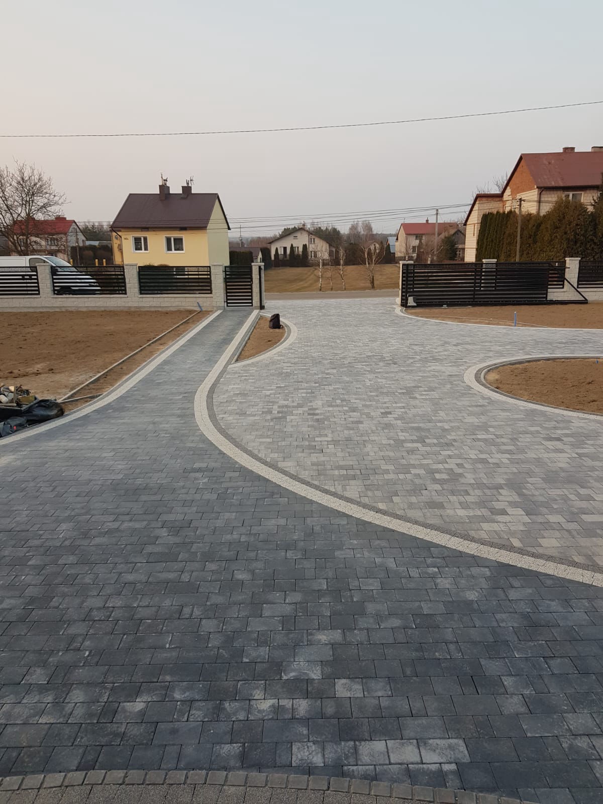 Ułożona szara kostka brukowa tworzy ścieżkę i podjazd, z jasnymi obrzeżami. W tle domy i ogrodzenia panelowe z bramami wjazdowymi. Widok z góry.