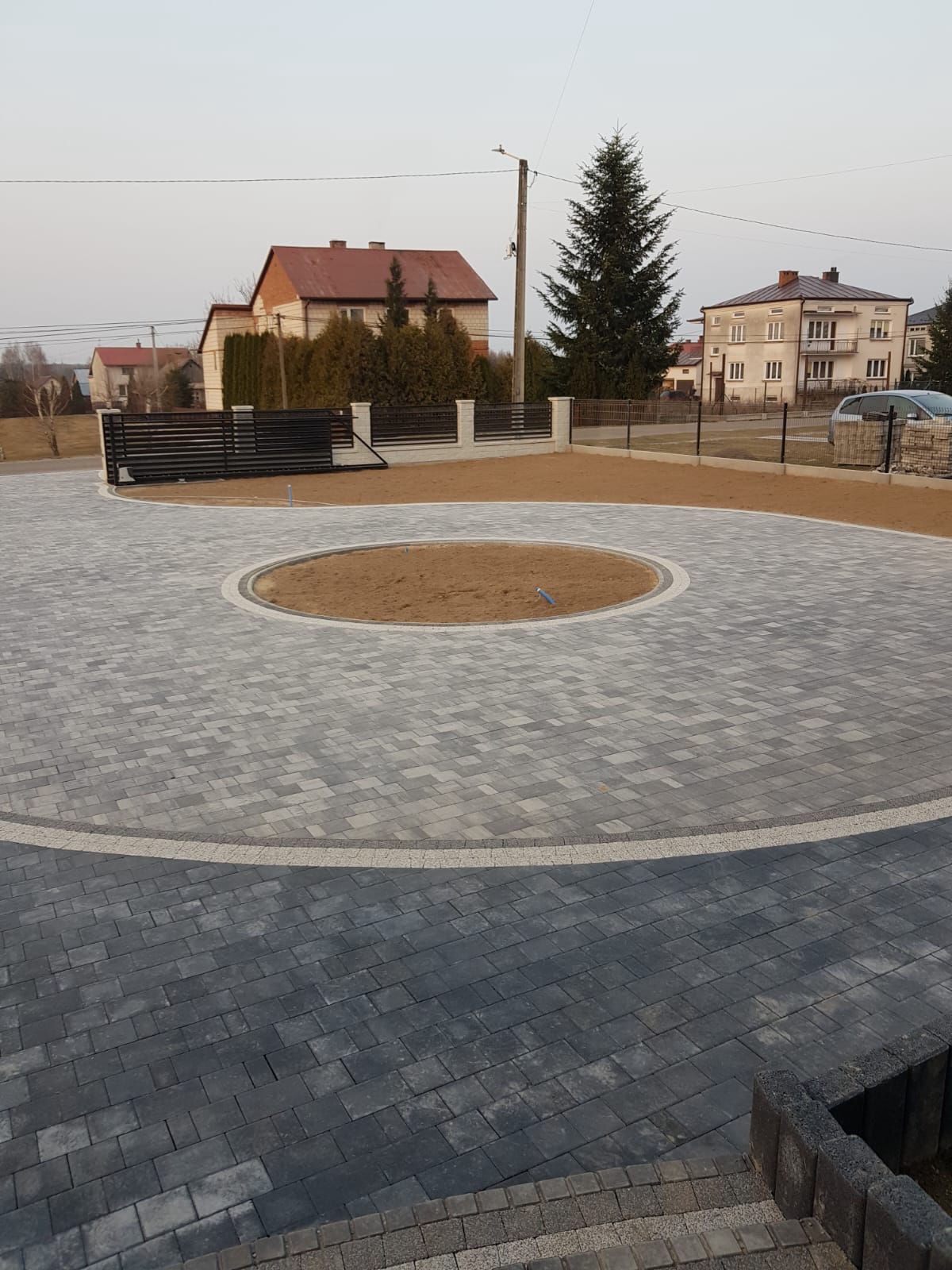 Wykończony podjazd z szarej kostki brukowej z okrągłą rabatą pośrodku, otoczony ogrodzeniem panelowym i bramą przesuwną. Widok na domy w tle.