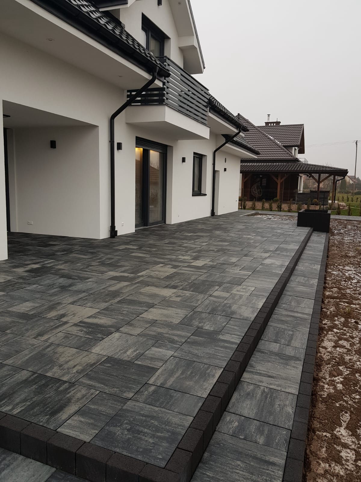 Nowoczesny taras z kostki brukowej w odcieniach szarości, przylegający do białego domu z czarnymi akcentami. Widoczna pergola w tle. Chłodna kolorystyka.