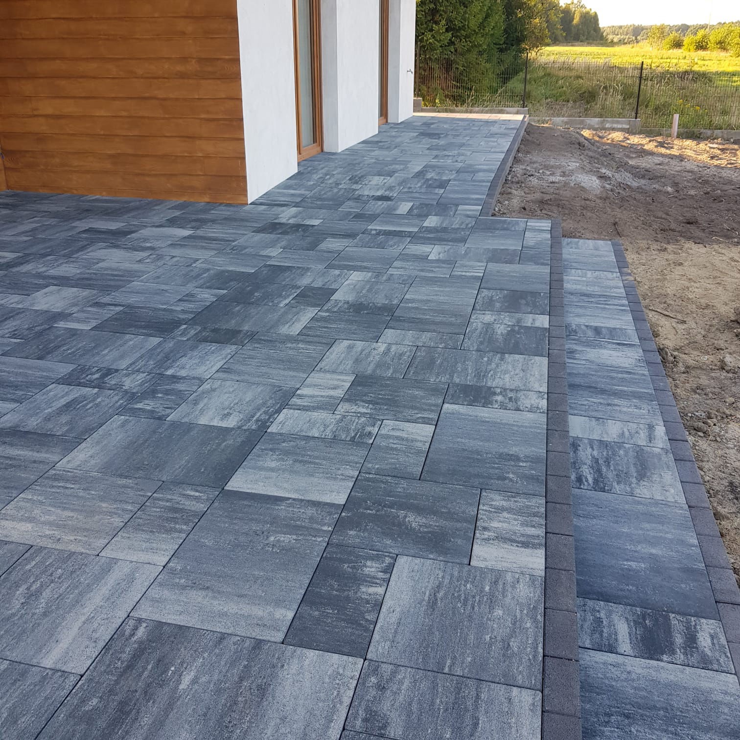 Nowoczesny taras z szarej kostki brukowej, z subtelnym wzorem ułożenia. Widoczne schody i fragment elewacji budynku z drewnianym wykończeniem.