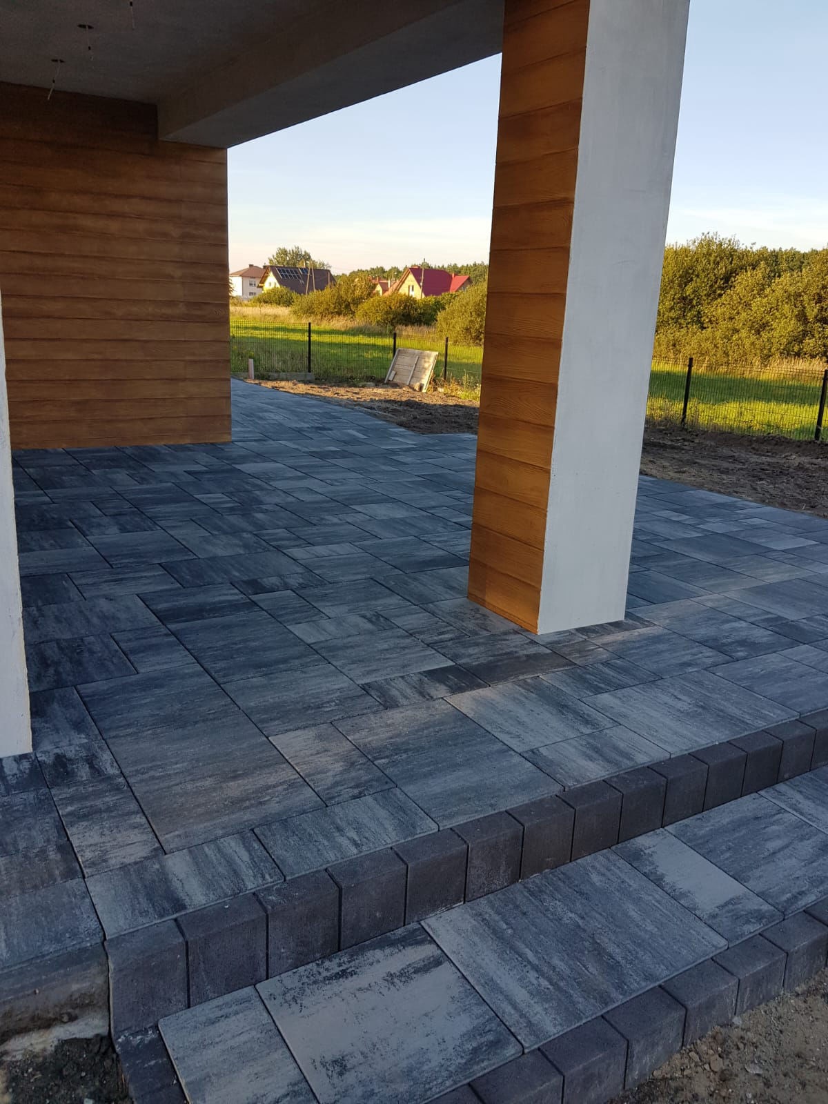 Nowoczesny taras z szarej kostki brukowej ułożonej w geometryczny wzór, widok na ogród i domy w tle, jasne oświetlenie.