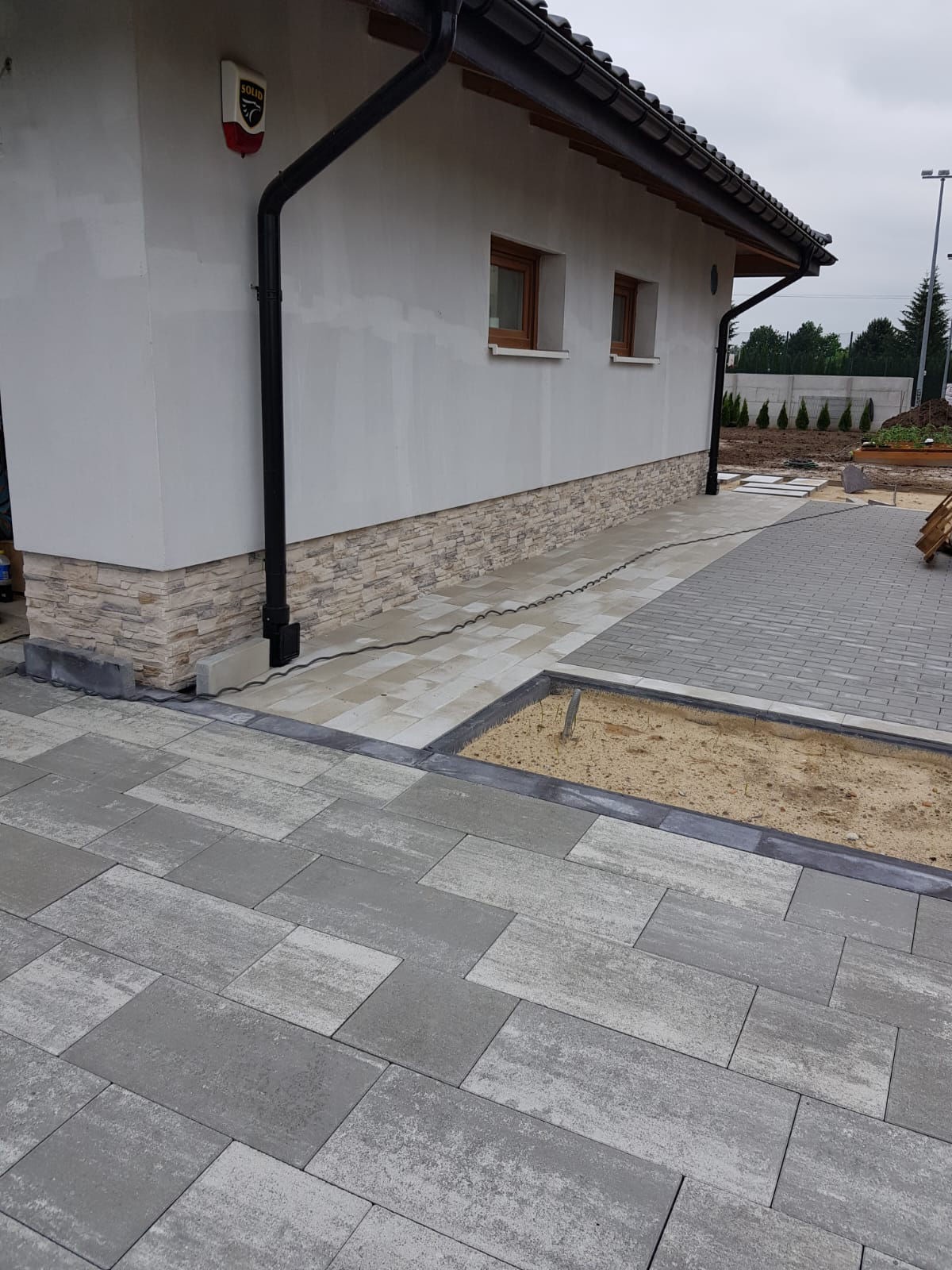 Szara kostka brukowa o nieregularnym kształcie, ułożona wokół rabaty z piaskiem, przylegająca do ściany budynku z brązowymi oknami i rynną.