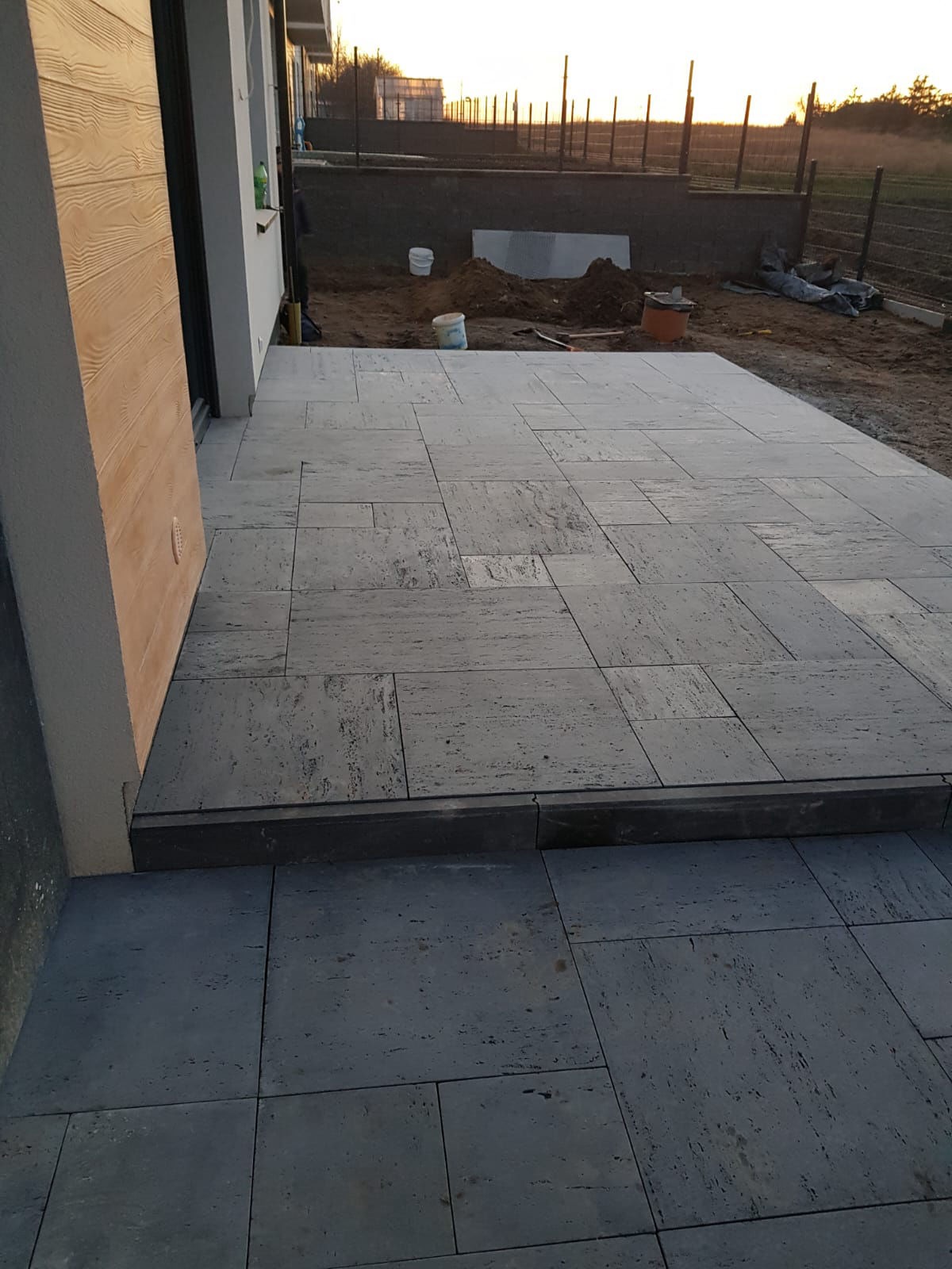 Ułożona kostka granitowa w odcieniach szarości, tworząca taras przy budynku. W tle widoczne prace ziemne i ogrodzenie.