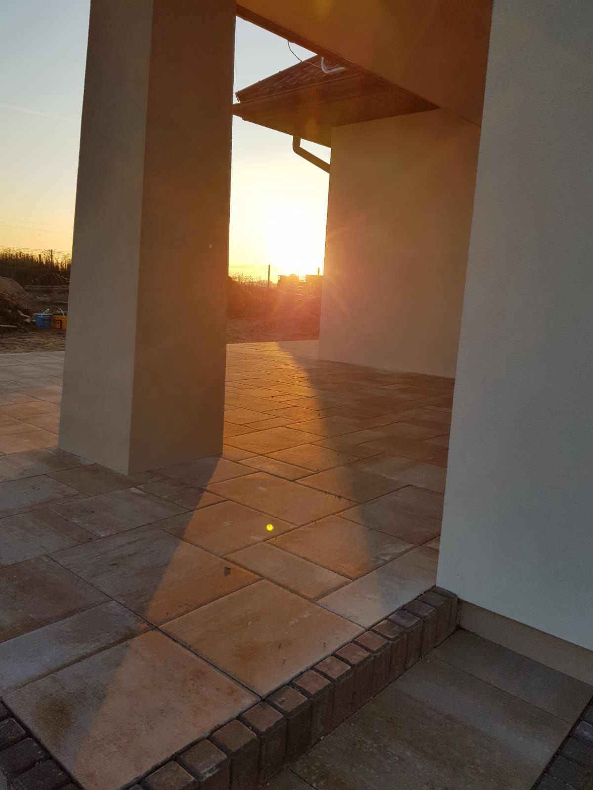 Ułożona kostka brukowa w odcieniach beżu na podjeździe domu, widok przez kolumny, w tle zachodzące słońce. Charakterystyczny wzór ułożenia.