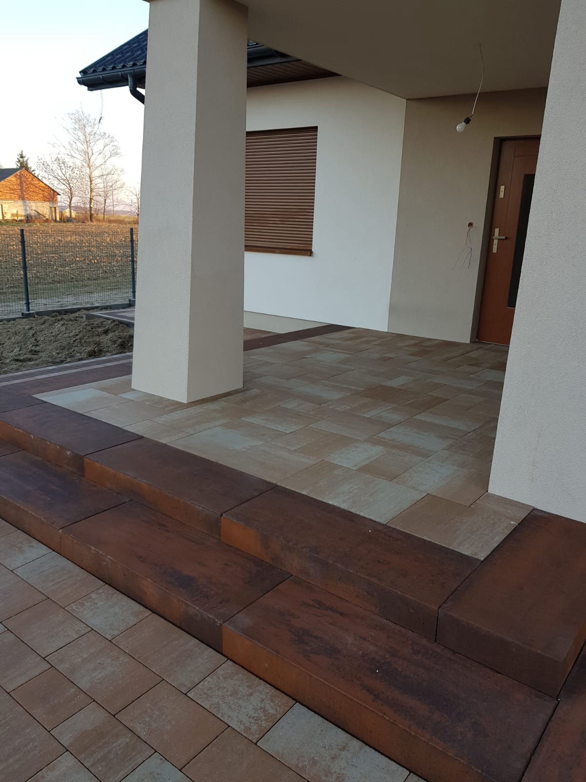 Wykończony taras z kostki brukowej w odcieniach brązu i beżu, z widocznymi schodami i kolumną podtrzymującą zadaszenie wejścia do domu.