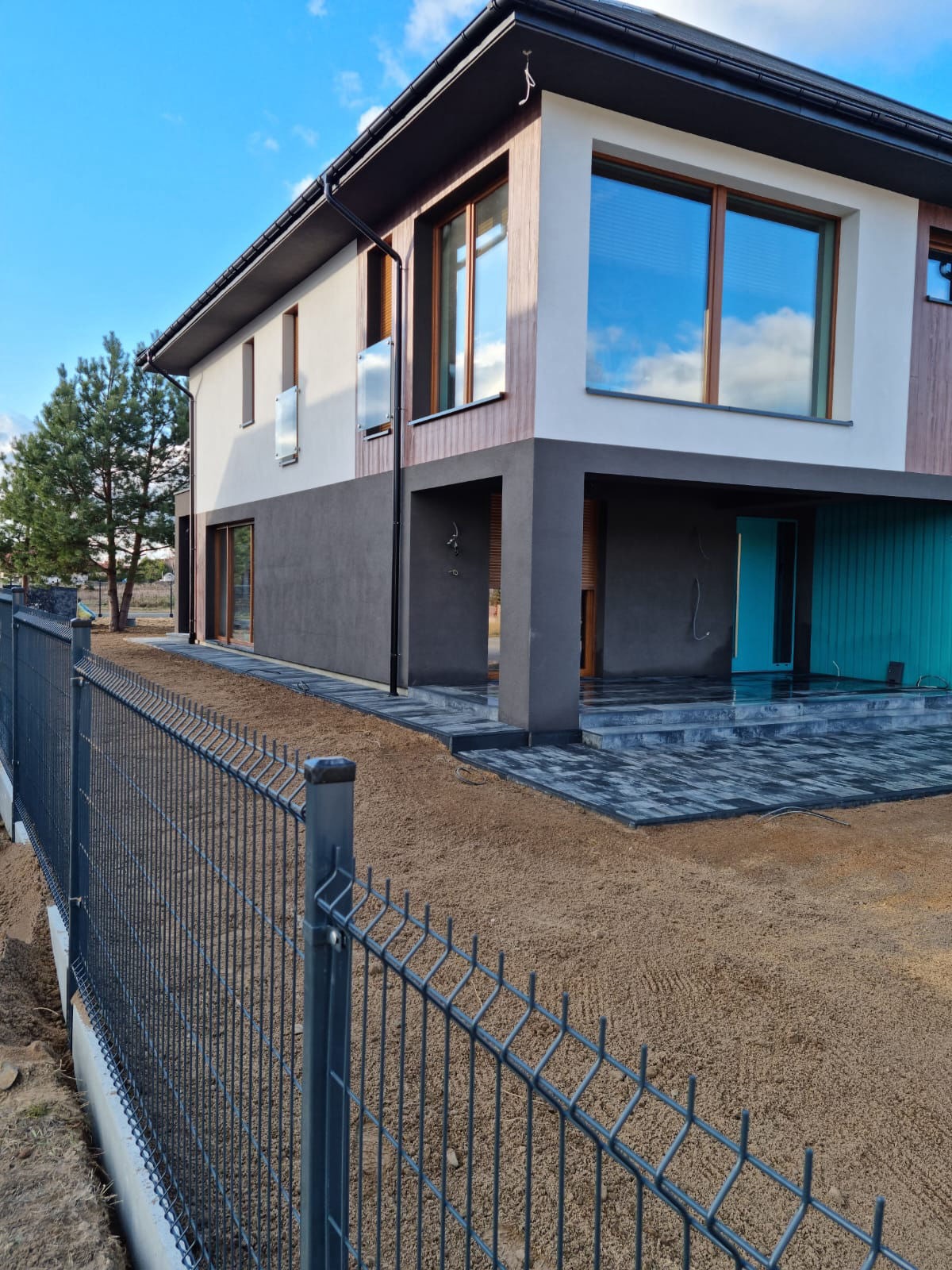 Nowoczesny dom z szarą kostką brukową i metalowym ogrodzeniem panelowym. Widoczny przygotowany teren pod dalsze prace.