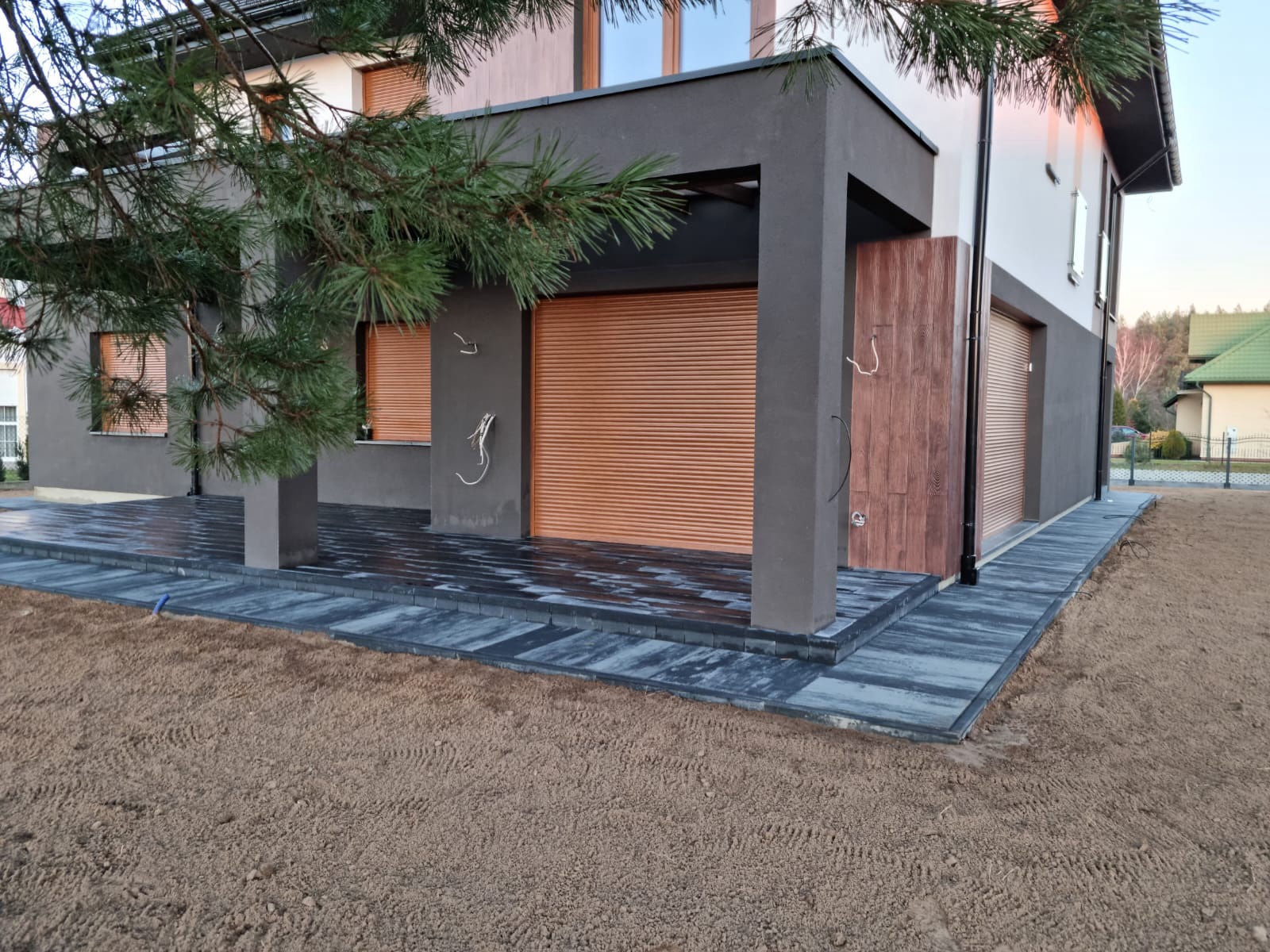 Elegancki taras z ciemnej kostki brukowej, dopasowany do nowoczesnej architektury domu z brązowymi roletami. Widoczne świeżo przygotowane podłoże.