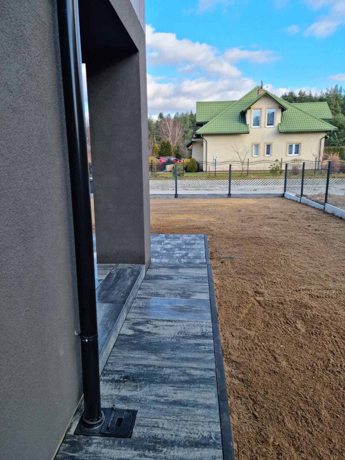 Nowoczesna kostka brukowa w odcieniach szarości prowadząca do domu. W tle ogrodzenie panelowe i dom z zielonym dachem, błękitne niebo. Widok z bliska na detale.