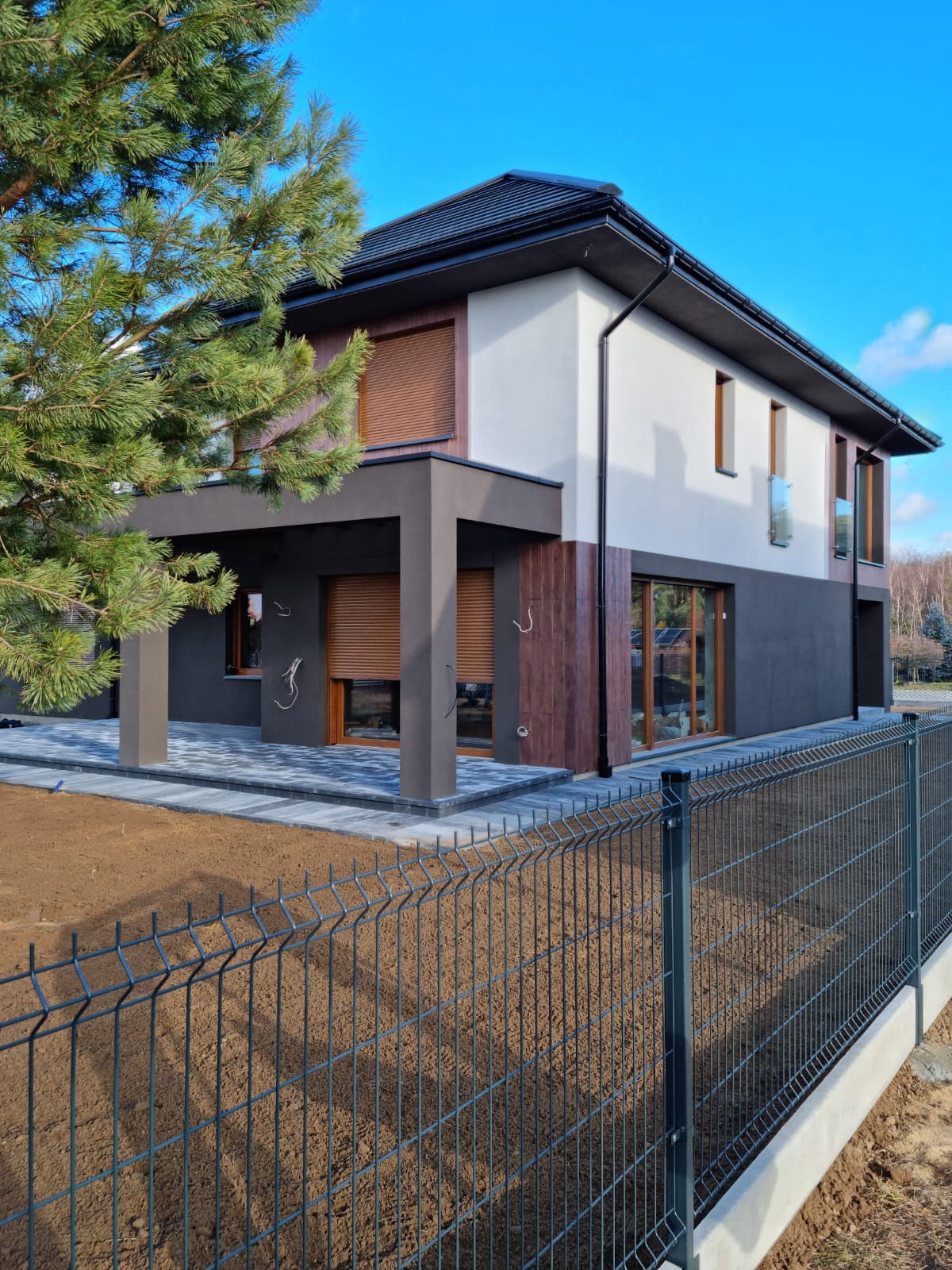 Nowoczesny dom z szarą kostką brukową i metalowym ogrodzeniem panelowym w Krakowie, widziany z niskiego kąta. Elementy drewniane na elewacji.