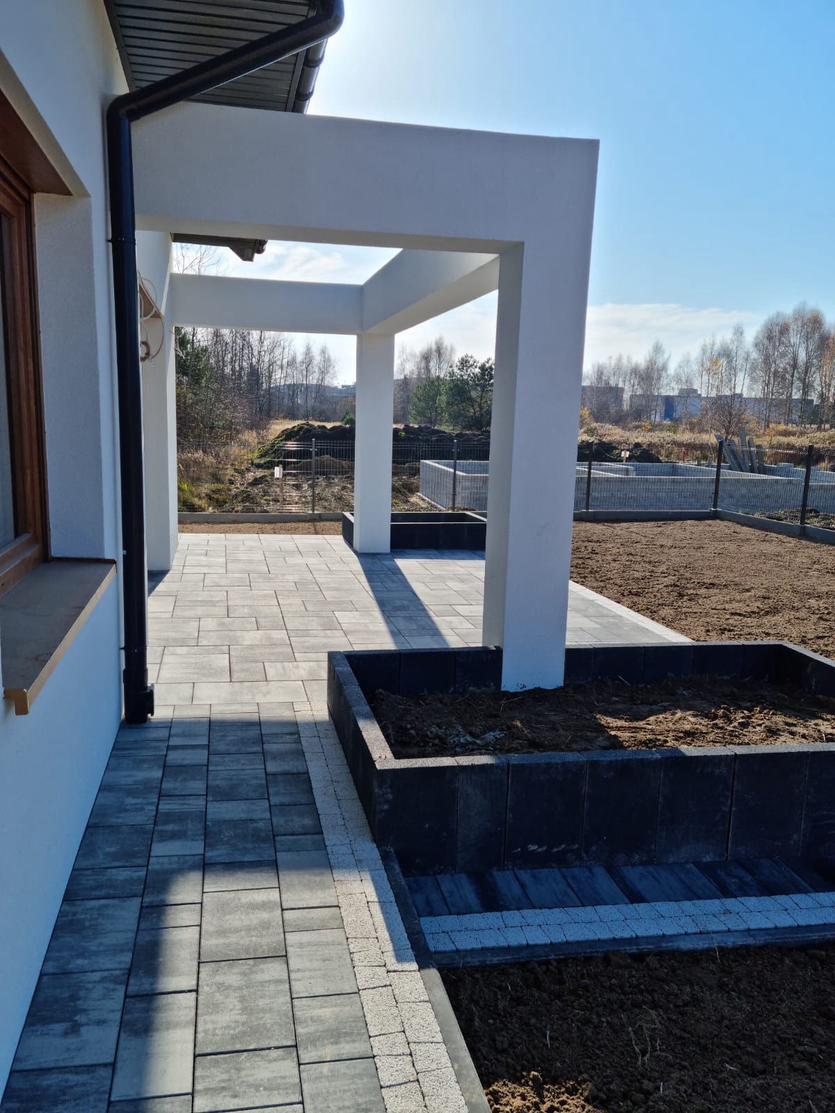 Ułożona kostka brukowa wzdłuż domu z białymi kolumnami, obok rabaty z czarnych bloczków. Widok na przygotowany teren pod ogród.