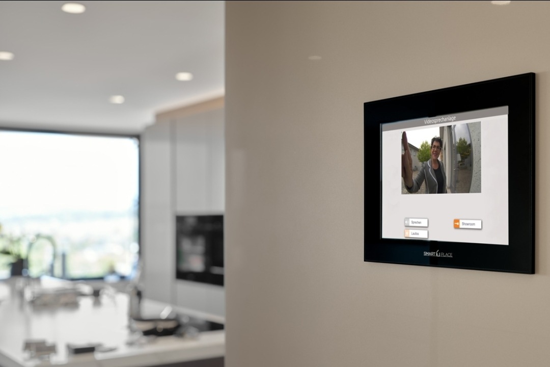 Panel wideodomofonu 'Smart Place' na ścianie, wyświetlający obraz osoby dzwoniącej. W tle widoczna nowoczesna kuchnia z oknem.