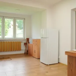 Jasne wnętrze kuchni z drewnianą podłogą i grzejnikiem. Widok na okno z zielenią, biała lodówka i fragment blatu z tkaniną.