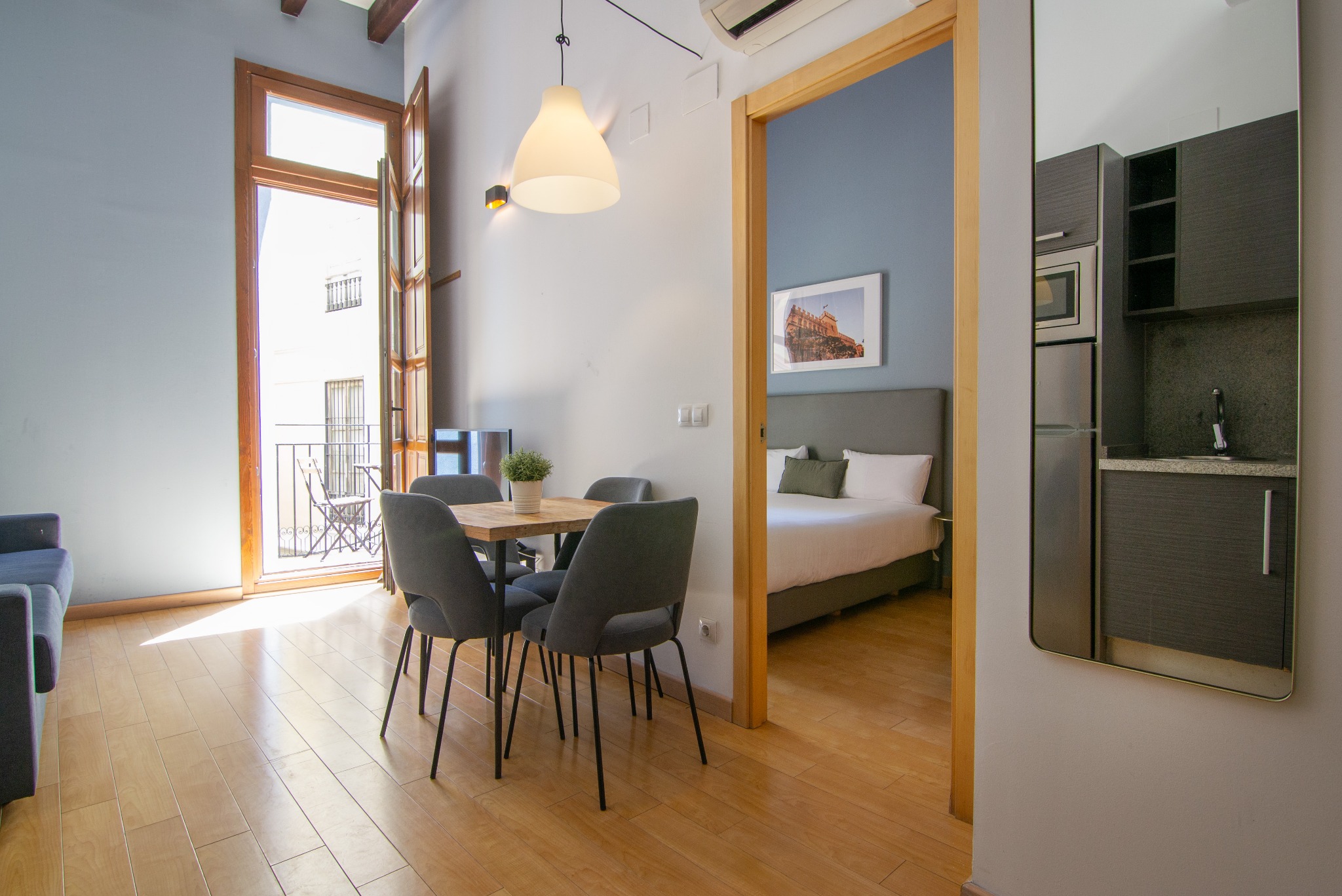 Jasne wnętrze apartamentu z balkonem, drewnianym stołem i krzesłami. Widok na sypialnię i aneks kuchenny odbity w lustrze. Naturalne światło.