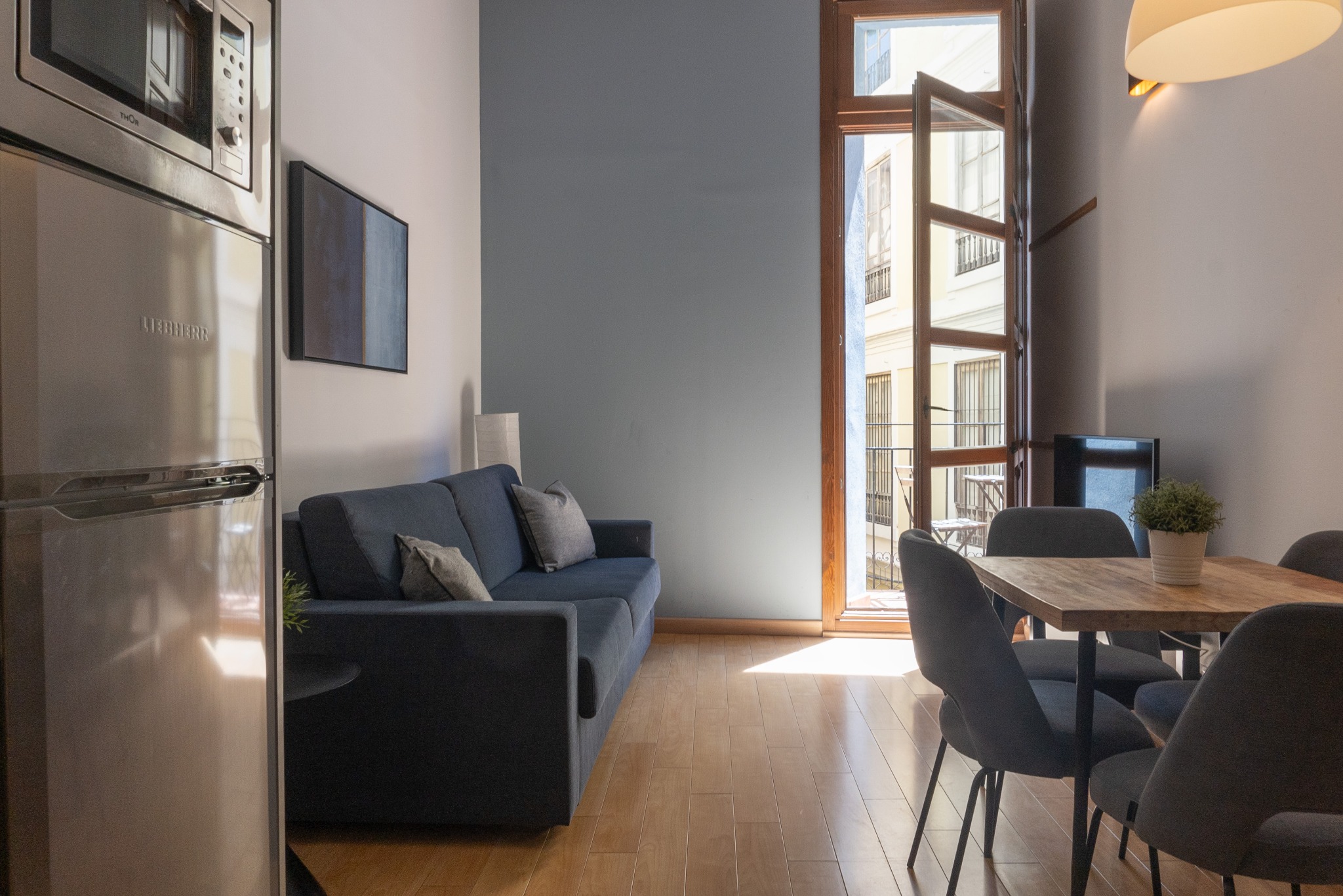 Jasne wnętrze mieszkania z drewnianą podłogą, niebieską sofą i stołem jadalnym przy otwartym oknie balkonowym, wpadające światło słoneczne.