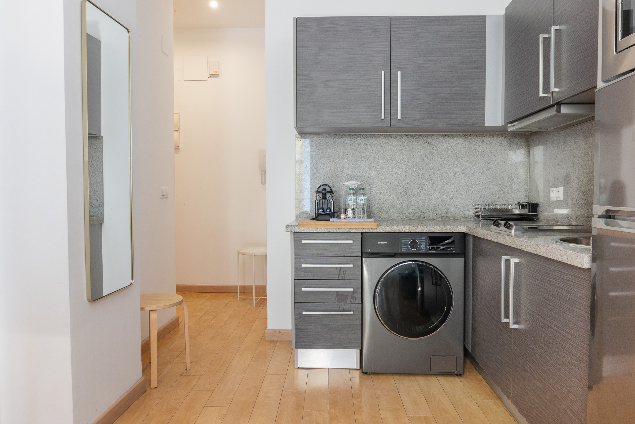 Nowoczesna kuchnia z pralką w zabudowie, szarymi szafkami i drewnianą podłogą. W tle lustro i krzesło. Minimalistyczny design z elementami wyposażenia.