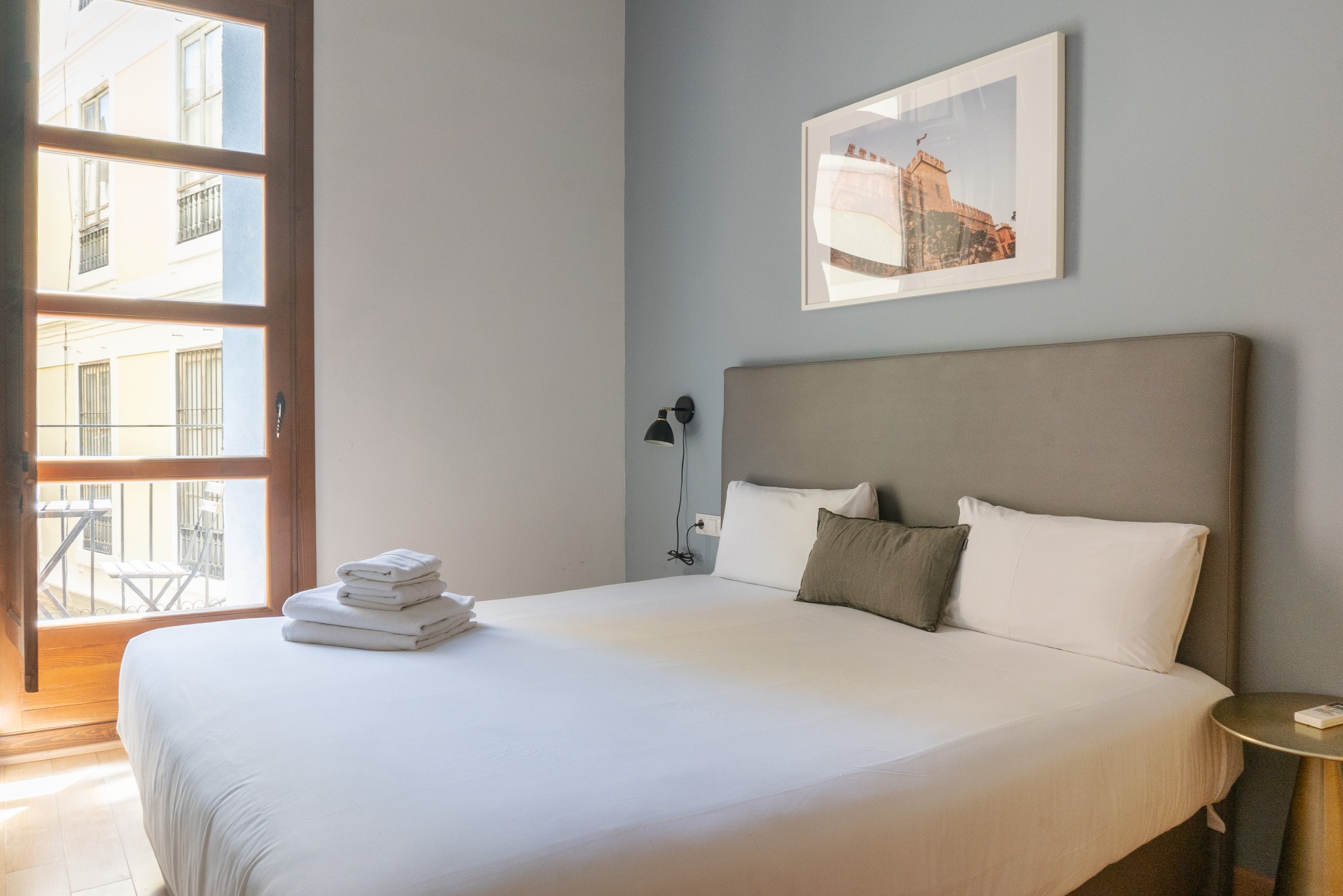 Jasny pokój hotelowy z balkonem: biała pościel, szare wezgłowie, drewniane okno z widokiem na balkon. Na ścianie obraz w ramie. Minimalistyczny design, spokojna atmosfera.