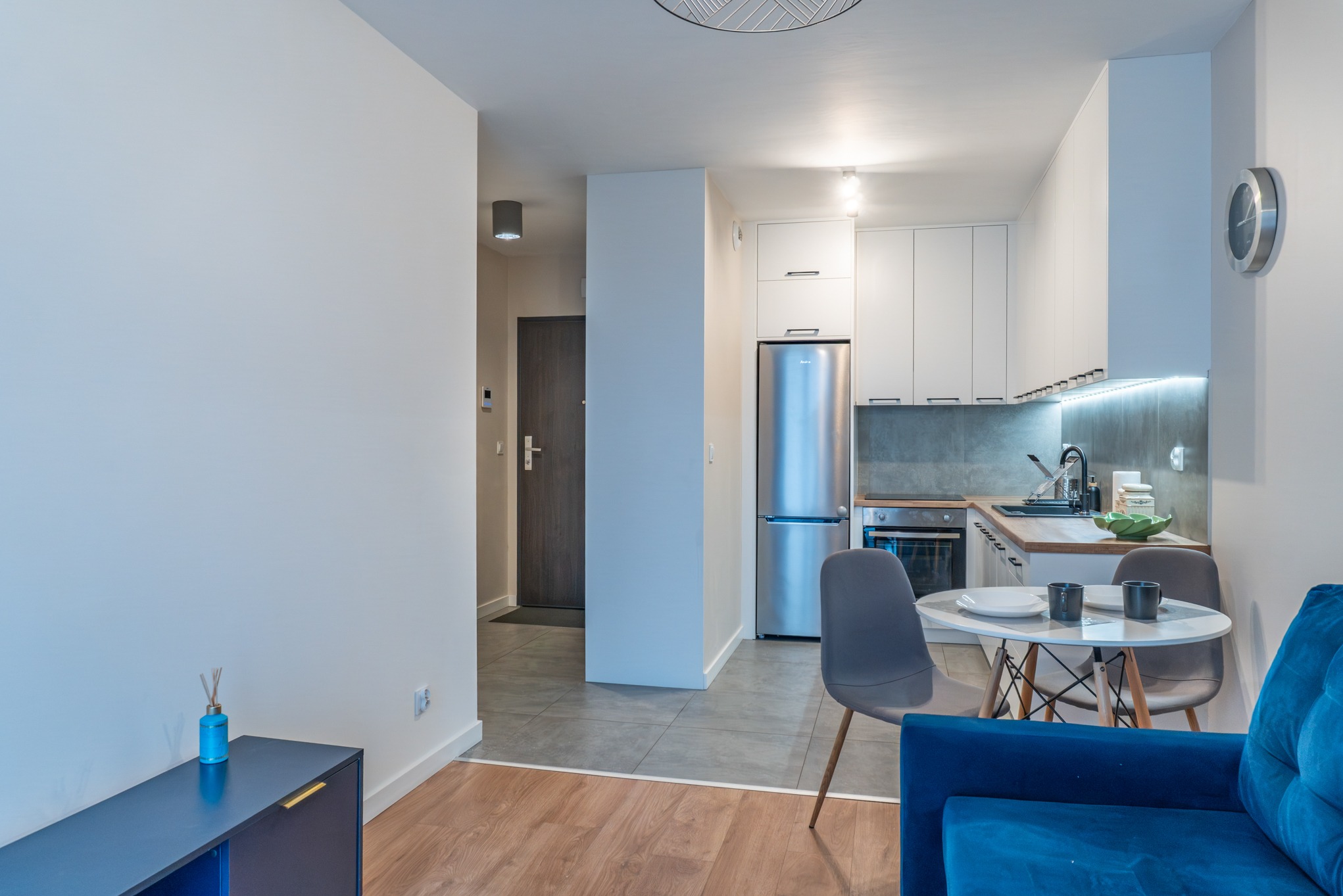 Nowoczesne wnętrze mieszkania: biała kuchnia z szarym blatem, okrągły stół z krzesłami, niebieska sofa i drewniana podłoga. Minimalistyczny design z elementami dekoracyjnymi.