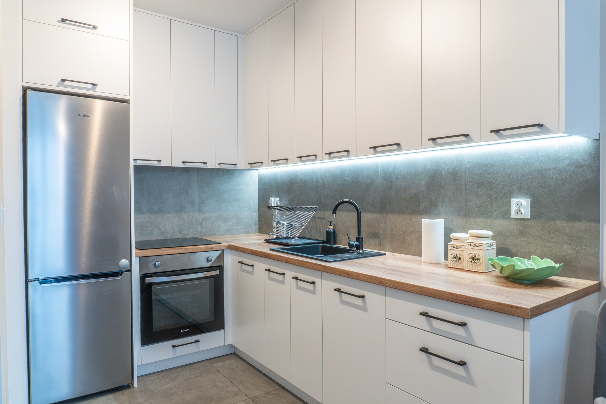 Nowoczesna kuchnia w bieli z drewnianym blatem, czarną armaturą i srebrną lodówką. Oświetlenie LED pod szafkami górnymi. Minimalistyczny design.