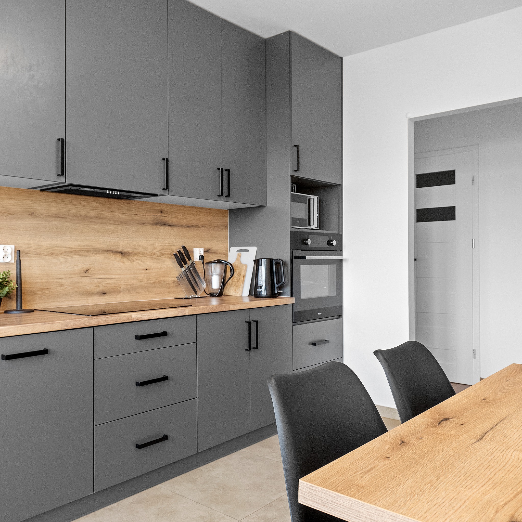 Nowoczesna kuchnia w szarościach z drewnianym blatem i czarnymi uchwytami, widok na stół i krzesła. Minimalistyczny design, oświetlenie punktowe.