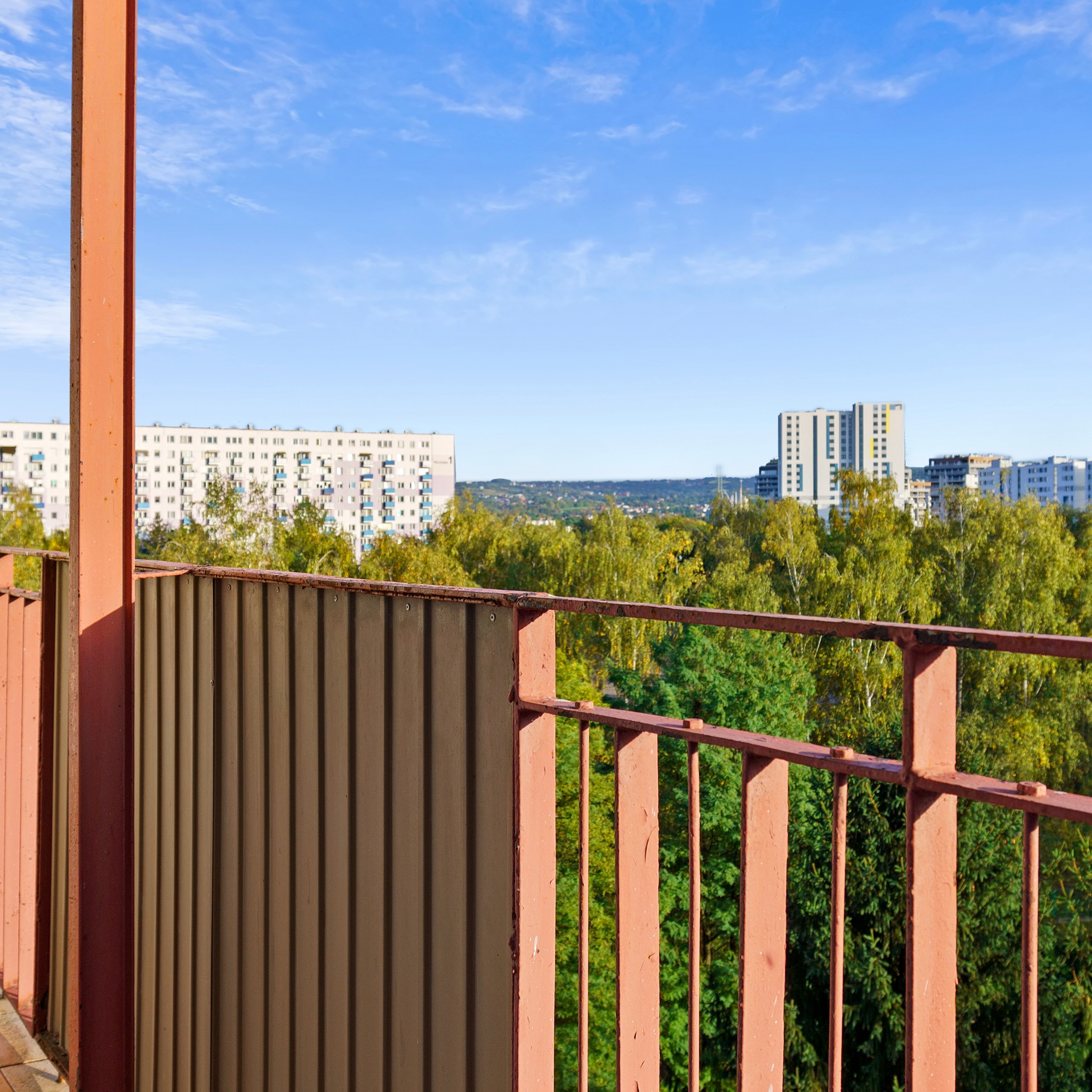 Widok z balkonu na osiedle mieszkaniowe i zieleń, z fragmentem metalowej balustrady w odcieniach brązu i pomarańczy na pierwszym planie, w słoneczny dzień.