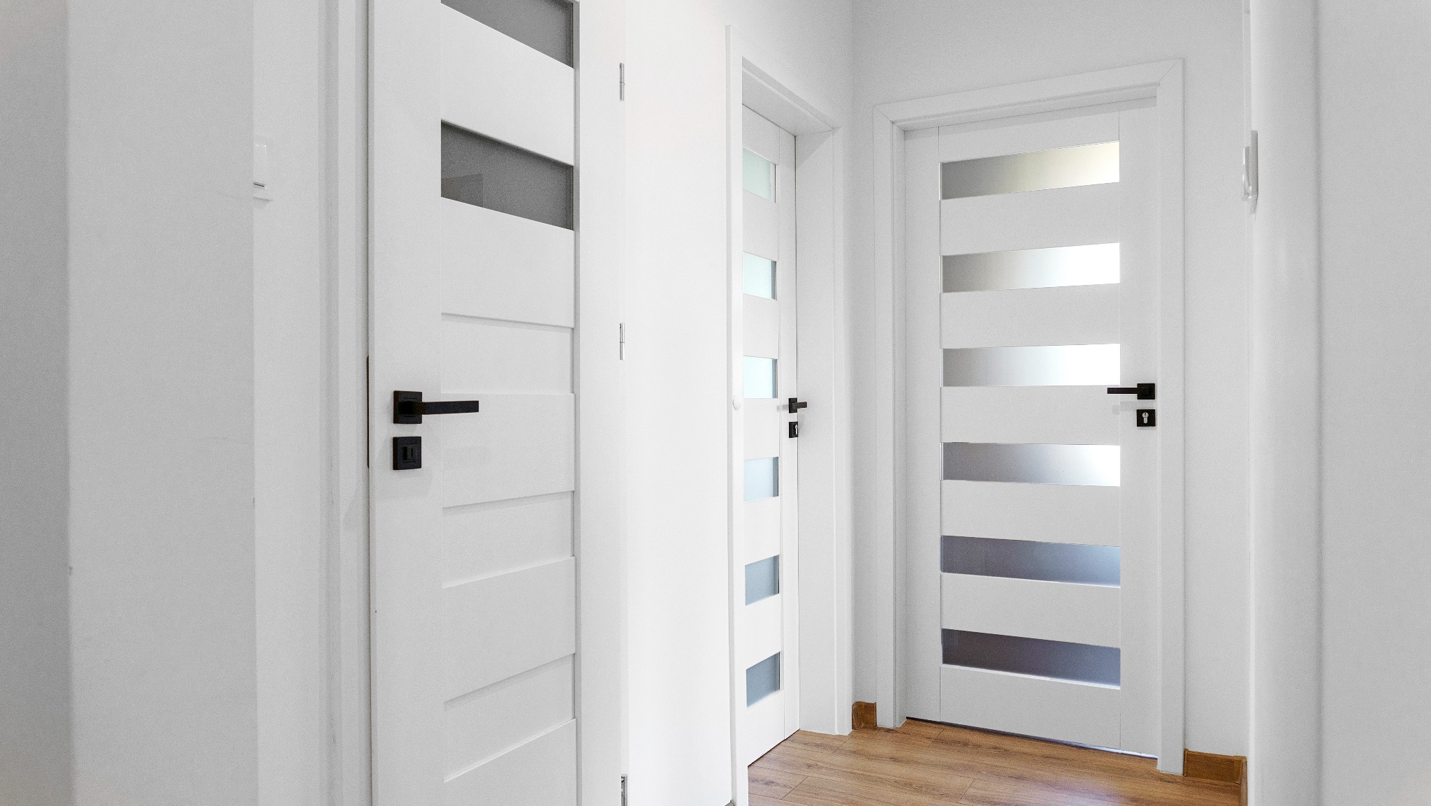 Białe drzwi wewnętrzne z czarnymi klamkami i przeszkleniami w minimalistycznym korytarzu z drewnianą podłogą. Nowoczesny design.