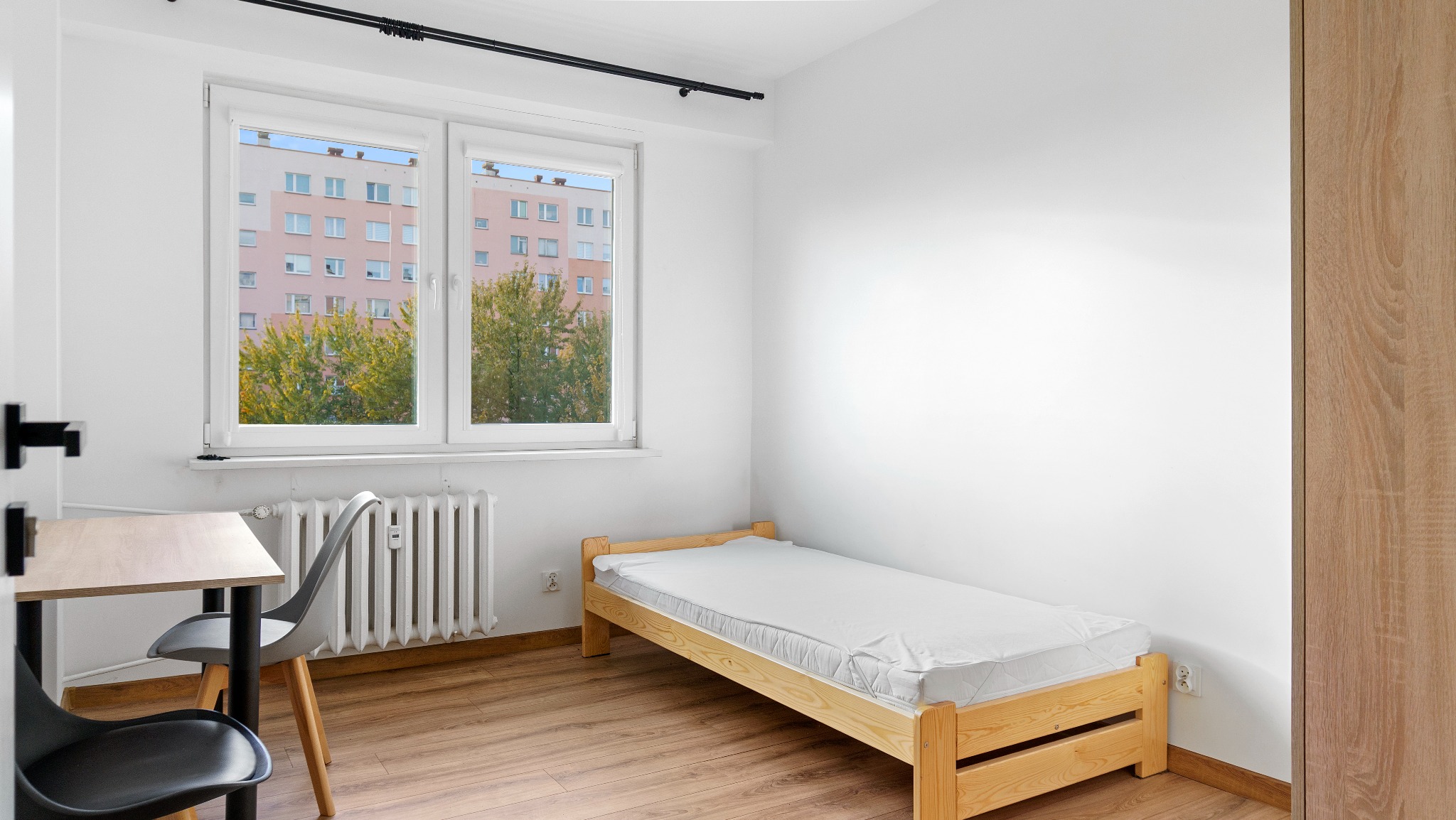 Jasne wnętrze pokoju z drewnianym łóżkiem, biurkiem i krzesłami. Za oknem widok na blok i drzewa. Minimalistyczny wystrój, parkiet na podłodze.
