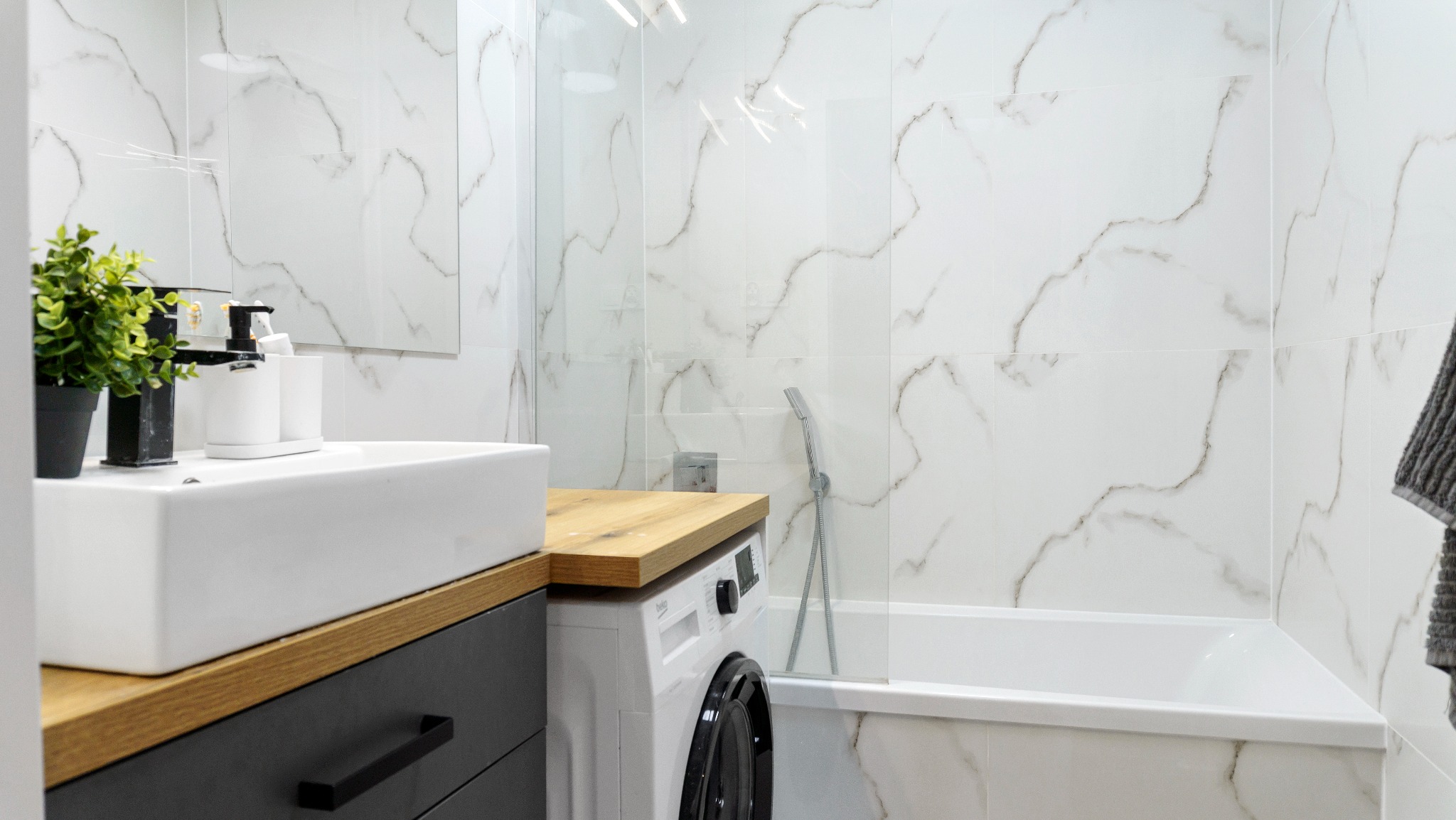 Nowoczesna łazienka z białymi płytkami imitującymi marmur, drewnianym blatem, białą umywalką i pralką. Kabina prysznicowa ze szklaną ścianką.